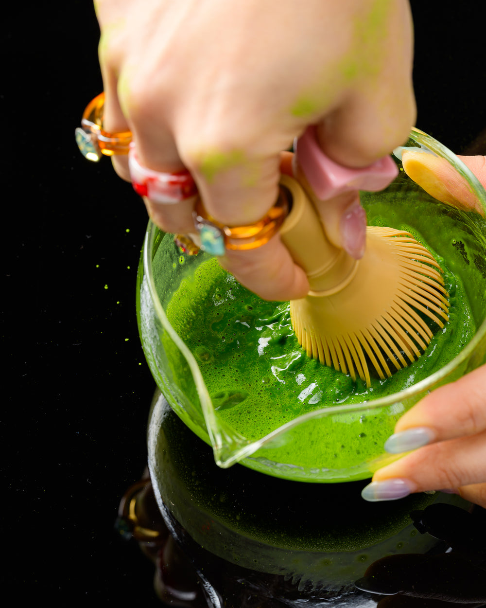 Hands whisking bright green matcha in a glass pitcher.