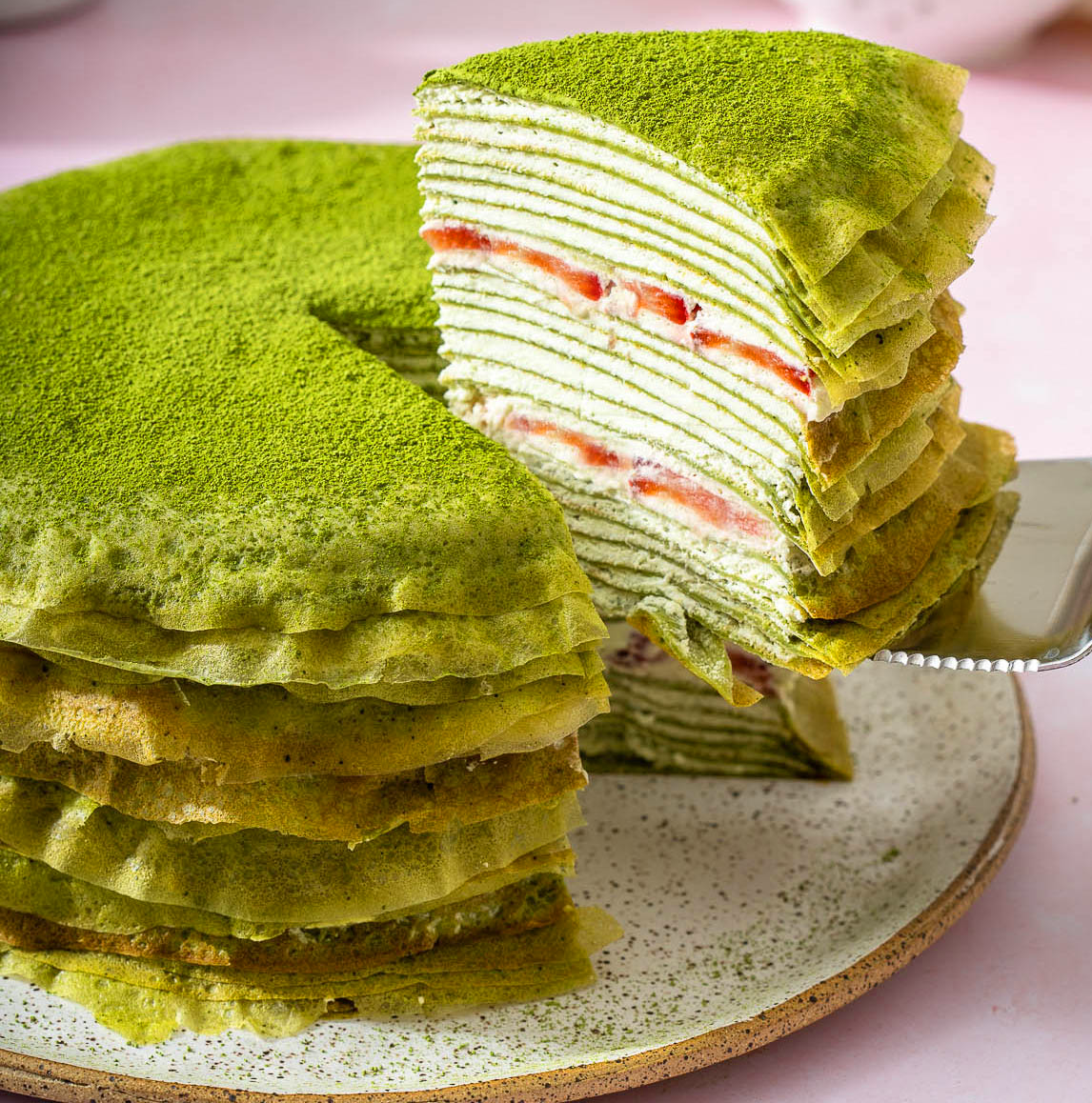 A slice of layered green matcha crepe cake filled with whipped cream and fresh strawberries being lifted from the cake on a ceramic plate, dusted with vibrant matcha powder.