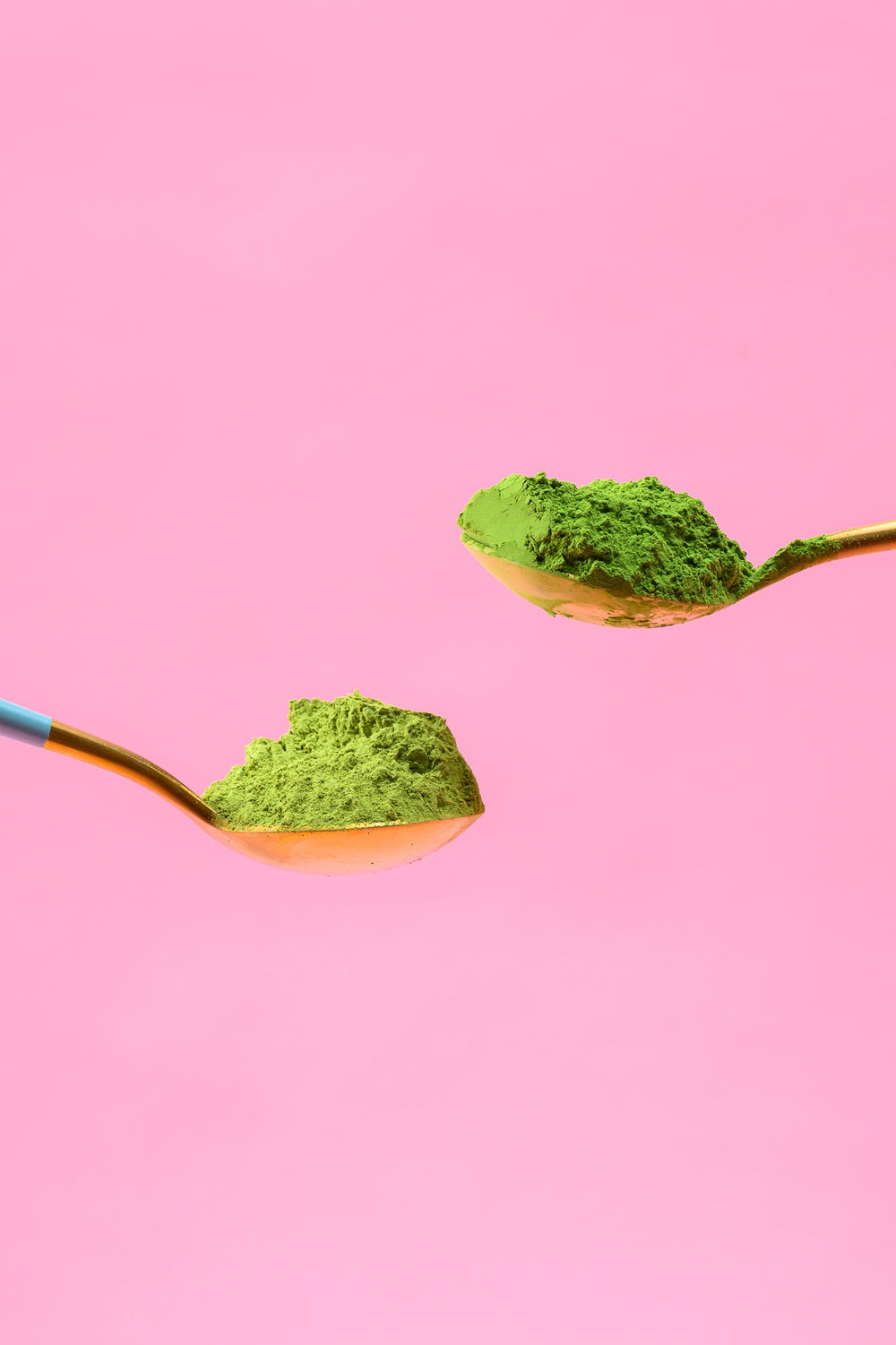 Green matcha powder on two gold-colored spoons against a pink background.