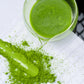 Top-down view of whisked ceremonial matcha tea in a clear glass spout pitcher, surrounded by vibrant green matcha powder spread across a white tiled background.