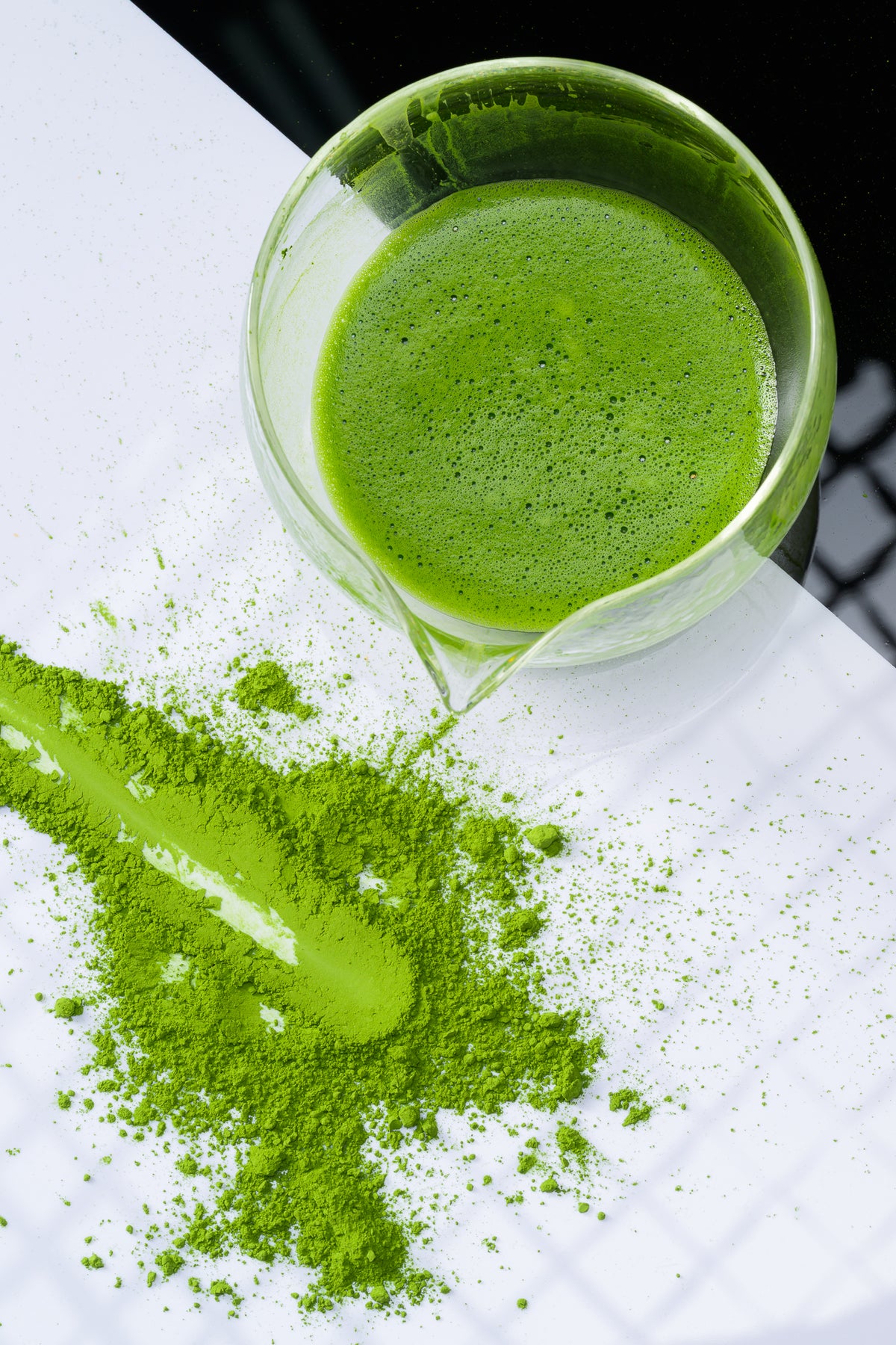 Top-down view of whisked ceremonial matcha tea in a clear glass spout pitcher, surrounded by vibrant green matcha powder spread across a white tiled background.